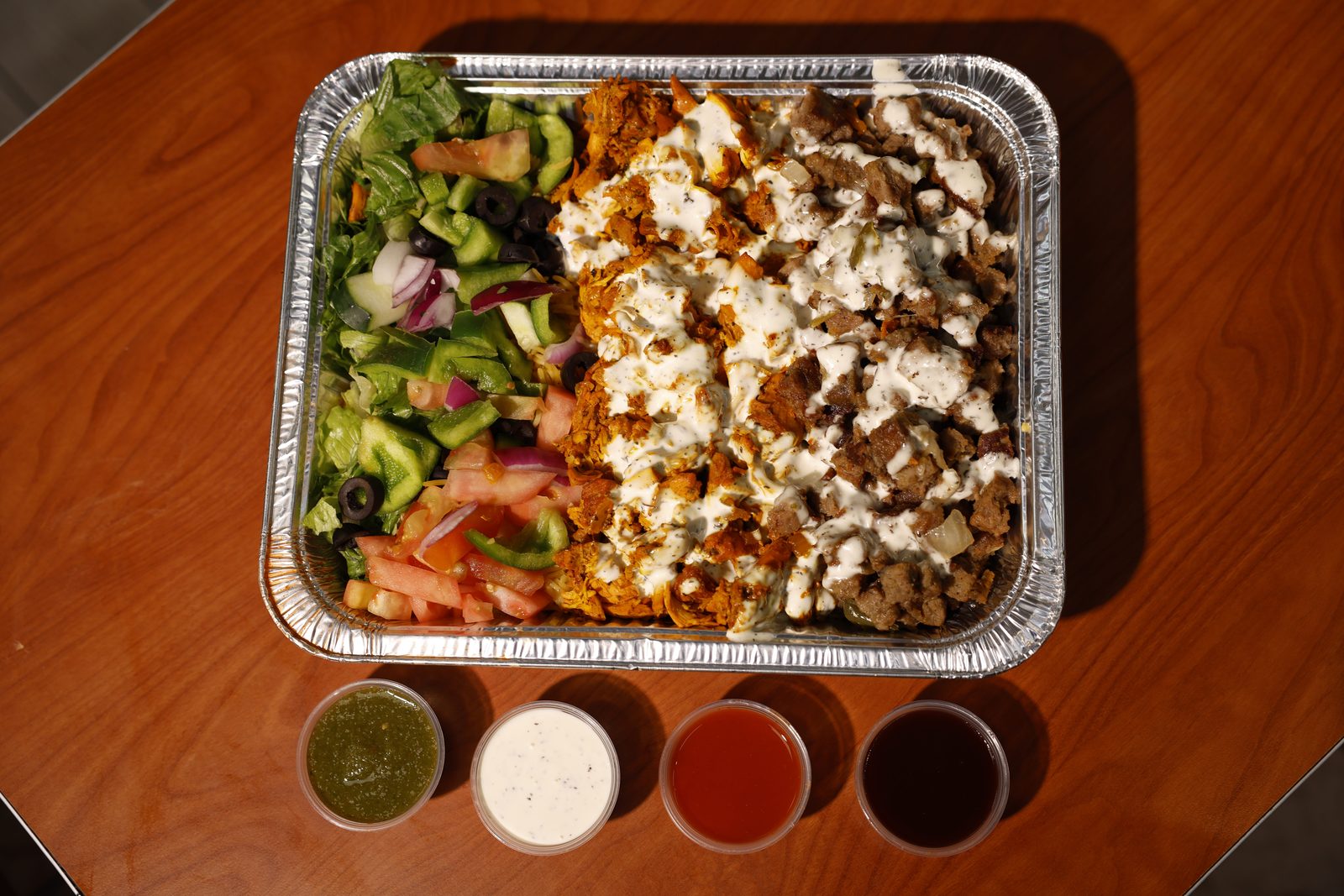 A Gyro King catering tray seen from above: chicken and gyro over rice with white sauce next to a fresh salad, with four sauce cups lined up on a wood table