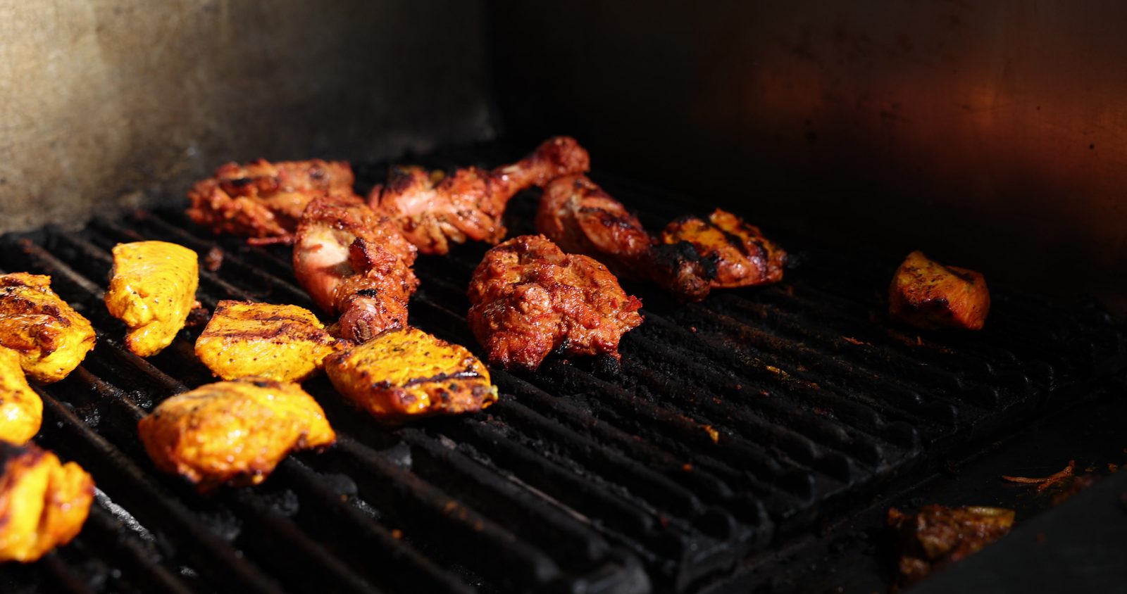 Chicken tikka pieces and marinated chicken legs charring on a black grill in the Gyro King kitchen