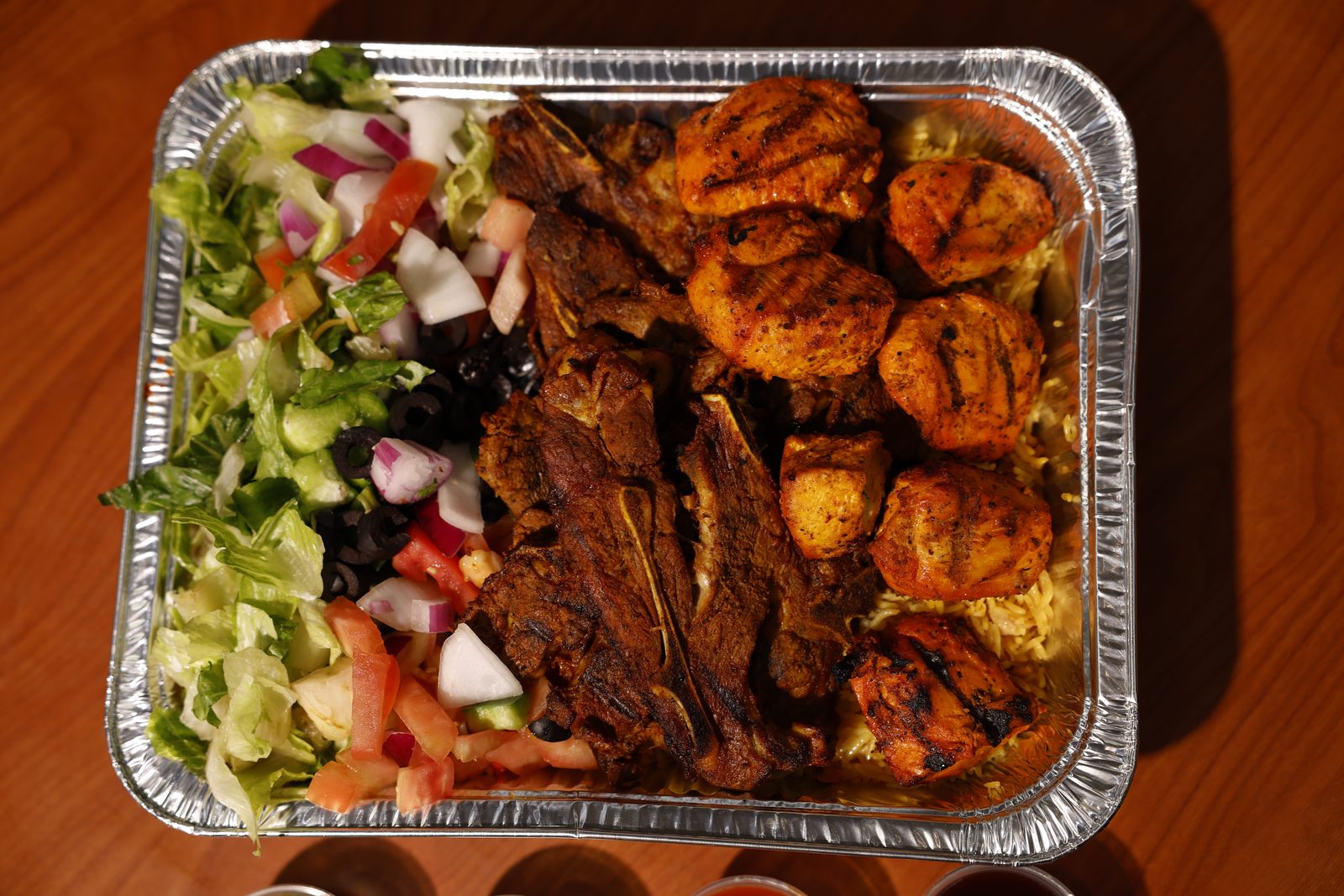 A Gyro King catering tray of lamb chops and grilled chicken tikka over rice, with chopped salad and olives, photographed from above on a wood table