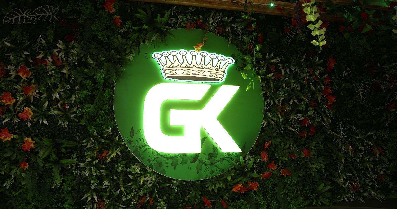 The glowing green GK crown logo sign on a wall of flowers and greenery inside Gyro King in Brooklyn