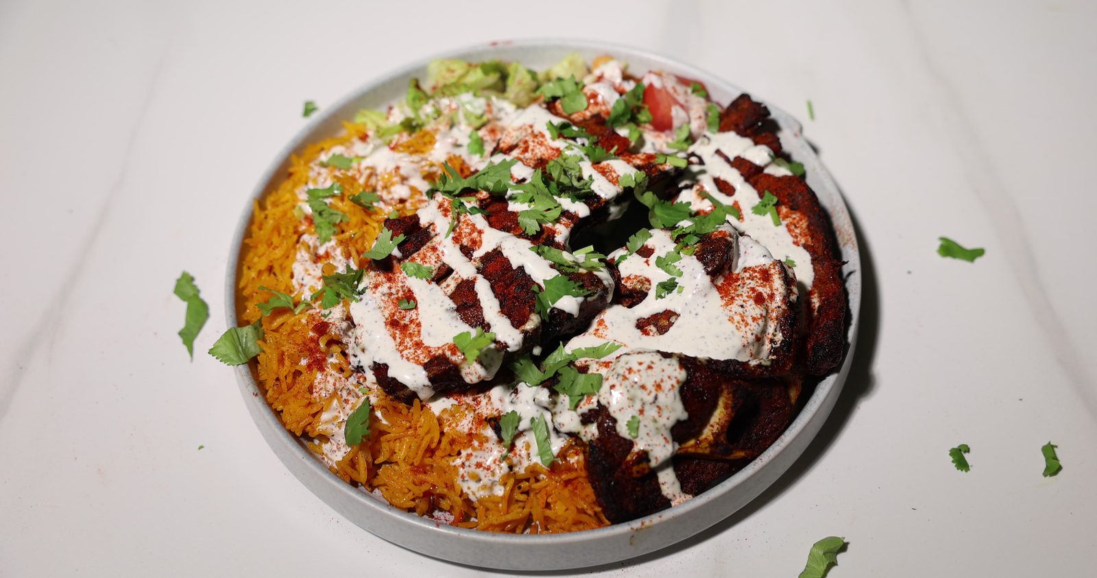 A plate of grilled chicken over yellow rice with white sauce and chopped herbs, with garnish scattered on a white marble surface, shot at Gyro King